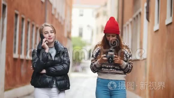 两个穿着温暖衣服的年轻女性旅行者走在狭窄的街道上。 一个女人在打电话，另一个