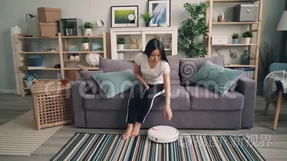 美丽的亚洲女孩打开机器人吸尘器，然后坐在沙发上，使用智能手机，享受方便