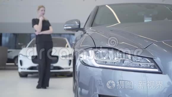 模糊的微笑的女人身材穿着黑色的衣服站在车展上环顾四周。 现代银色轿车的前照灯