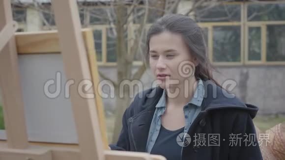 肖像成功，美丽的年轻女孩画家坐在木制画架前环顾四周。 女性艺术家