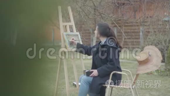 相当集中的年轻女画家坐在木制画架前画一幅画。 休闲女艺术家