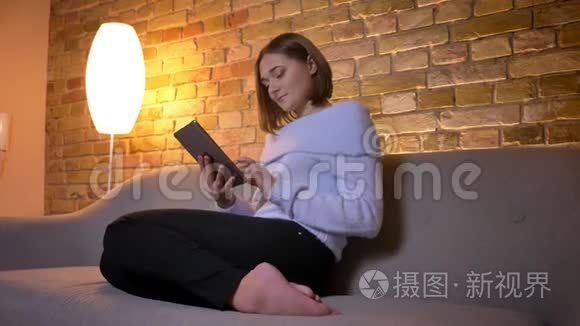 年轻漂亮的白人女性轻敲平板电脑，然后转向镜头，微笑着坐在