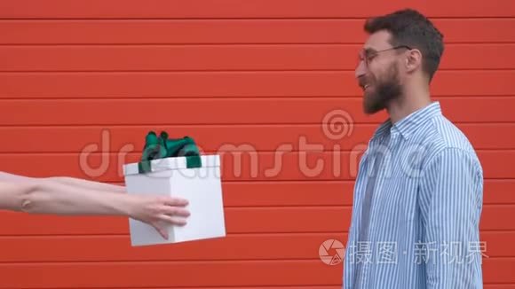 大胡子男子高兴地得到白色礼品盒与绿色蝴蝶结从女性手上红墙