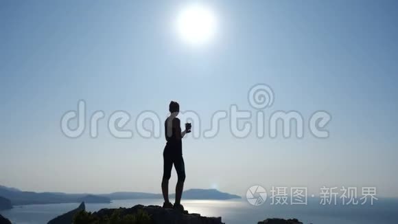 一位快乐的徒步旅行者女士的剪影，她端着一杯茶站在山顶上，在一个夜晚欣赏着美妙的景色