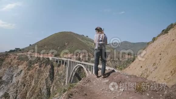 年轻的旅游妇女带着背包，在阳光明媚的加州大苏尔的比克斯比峡谷桥景点观看史诗般的风景。