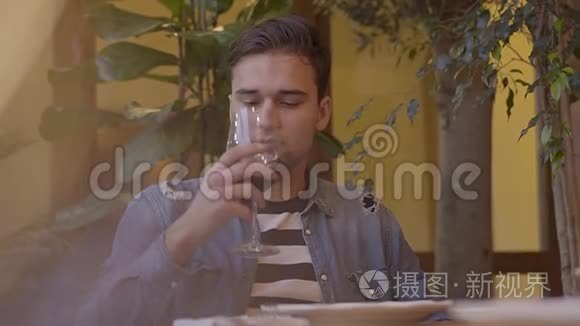 一幅迷人的家伙喝着酒，酒杯坐在餐馆的桌子旁。 年轻人一边享受