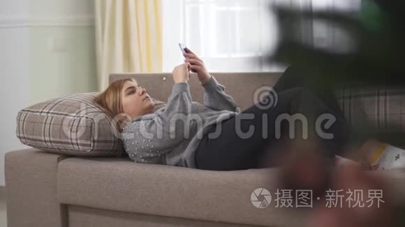 胖乎乎的年轻女人在室内打手机，躺在大窗户附近的沙发上。 可爱加上大小的女性放松