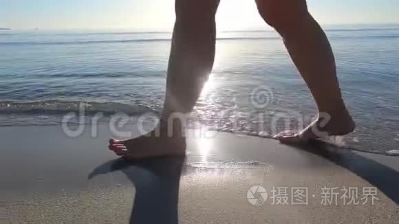 一个年轻女人的腿沿着海岸走着，海浪拍打着她的脚，在黎明时的背光下踩着沙子。