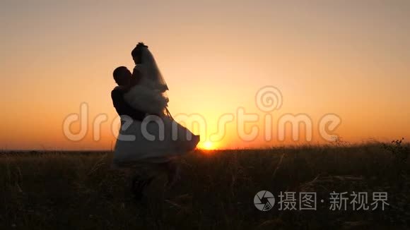情人新娘穿着白色礼服，新郎在夏天的夕阳下在田野里跳舞。 年轻的已婚夫妇亲吻拥抱