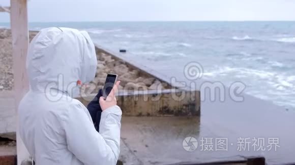 穿白色夹克的女人拿着手机大风浪站在堤岸上。 后景。