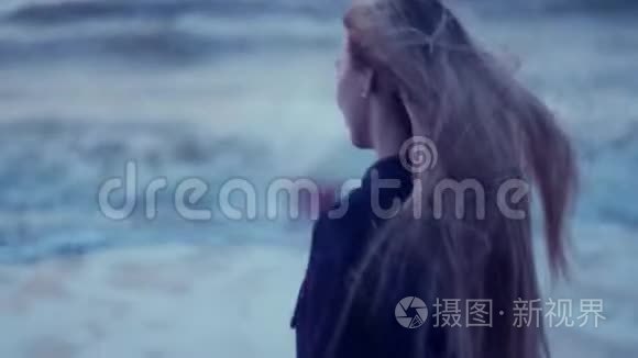 美丽的女孩看着大海，转向相机，她的头发在背景下发展，风，波浪