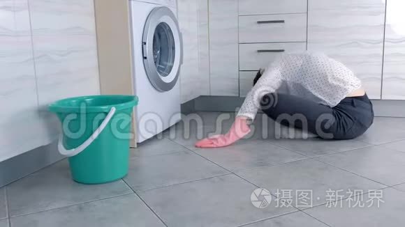 穿着粉红色橡胶手套的女人用布洗厨房家具。 坐在地板上。 侧视。