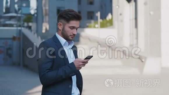 年轻英俊的商人站在大办公楼附近。 在他的智能手机上输入信息。 穿着