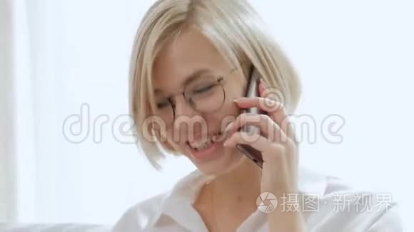一个年轻漂亮的金发女人戴着眼镜坐在沙发上用手机说话