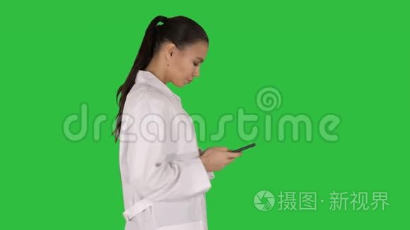 开朗可爱的年轻女性医生在绿色屏幕上行走时，在手机上键入文字，Chroma键。