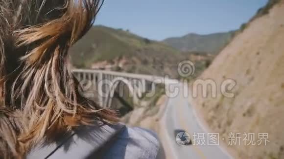 后景年轻的旅游妇女带着背包和飞扬的头发，在比克斯比峡谷桥点，大苏尔观看史诗般的风景。