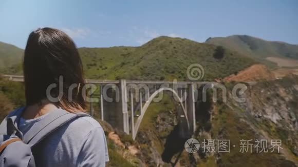 后景年轻兴奋的旅游妇女与背包观看史诗般的风景标志性比克斯比峡谷桥，大苏尔美国。