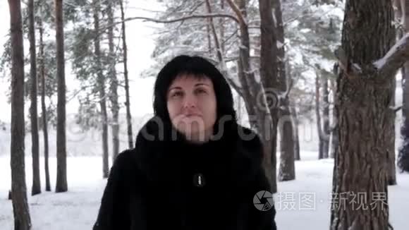 一个穿着毛皮外套的年轻黑发女人在霜冻的日子穿过一片雪林或公园，看着镜头。 照相机