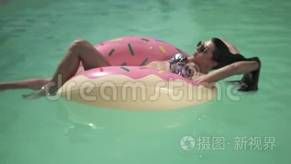 戴着太阳镜的漂亮年轻女人喜欢在游泳池里游泳，躺在粉红色的橡胶圈上。 孤独的女士的悠闲