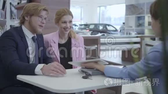 穿着时尚西装的汽车经销商在平板电脑上向年轻的成功夫妇展示他们的新车的信息。 汽车