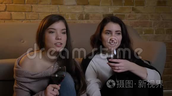 年轻的白种人女性朋友坐在地板上，端着酒杯，在舒适的家里看电影