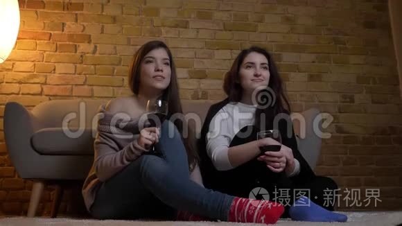 快乐的年轻白种人女性朋友坐在地板上，端着酒杯，在舒适的家里看电影