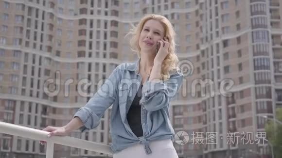 自信微笑的金发女人穿着牛仔裤夹克，在摩天大楼前用手机说话。 城市生活方式。 女性城市