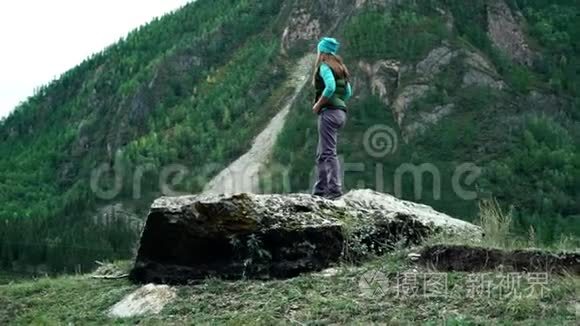 穿着背心和帽子的女旅行者站在树木覆盖的山上的一块大石头上