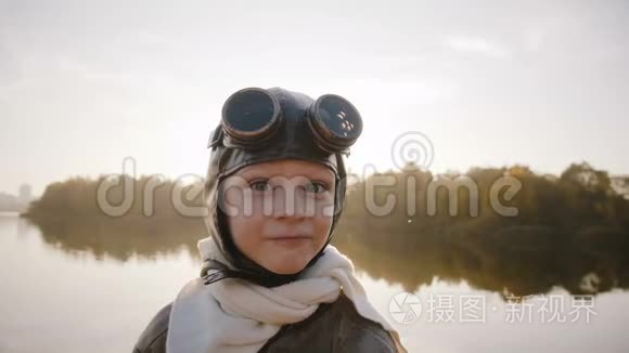 小男孩穿着老式的飞行员服装，戴着围巾和眼镜，看着相机，在湖边做着滑稽的表情。