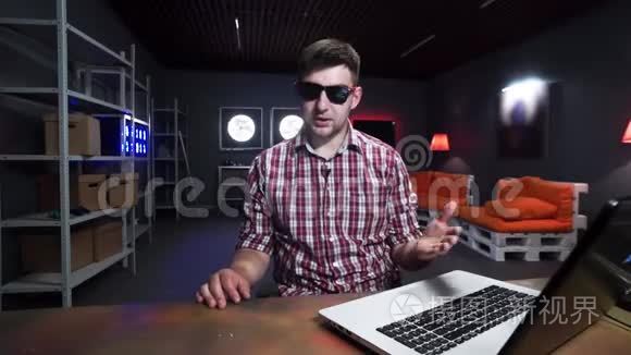 有趣的留胡子的家伙坐在桌子后面，在黑暗的房间里拿着笔记本电脑