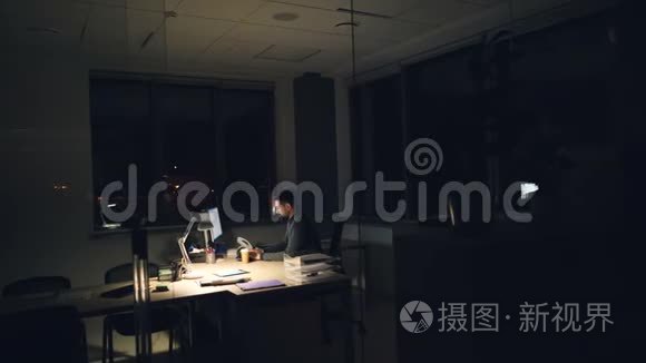 穿西装打领带的疲倦商人在办公室里用电脑工作，晚上坐在黑暗的房间里的工作台上