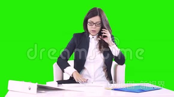 年轻的深色头发的年轻女人坐在办公室的办公桌后面打电话。 桌子上有文件