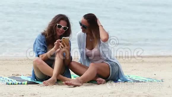 阳光明媚的一天，两个年轻的女人正在海滩上用手机自拍。