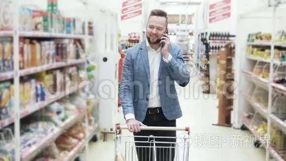 穿着白色衬衫的年轻微笑男子正在智能手机上交谈，并在超市的货架之间行走着商品。