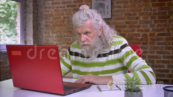 可爱的白种人老人坐在他的电脑旁边，写着笔记，白发和胡须，随意而寒冷