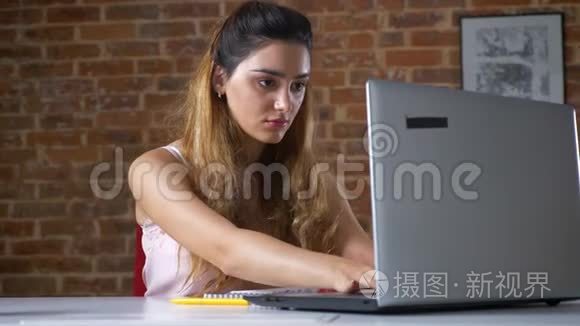 在红砖工作室里，一个正在用笔记本电脑工作的白种人女孩疲惫地看着她，并为放松而停下来