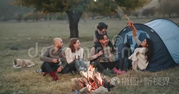 美丽的年轻家庭和他们的朋友在野餐时坐在篝火旁，爸爸和妈妈在玩木头