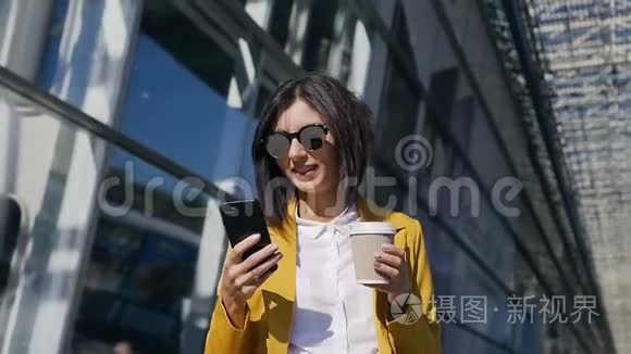 年轻的戴墨镜的职业女商人走在城市的街道上，喝着咖啡，用现代智能手机-