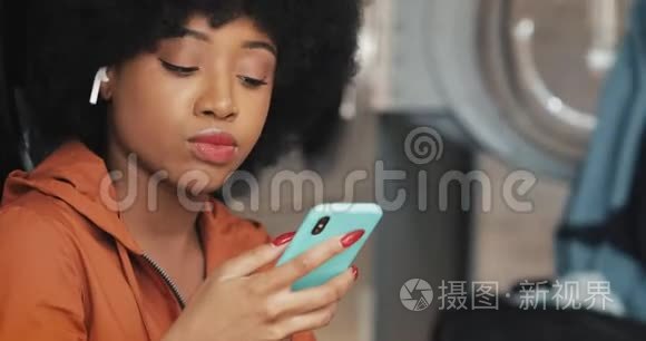 微笑着年轻的非洲裔美国妇女使用智能手机在自助洗衣店。 她听音乐唱歌。 自助服务公众