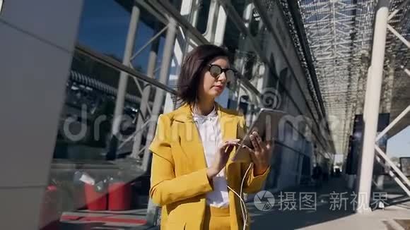 一位戴着耳机使用平板电脑的年轻漂亮女商人站在商业中心的城市街道上