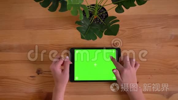 在木桌和植物背景上水平平板的绿色屏幕上打字的女子手的顶部镜头。