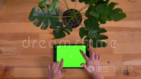 在木桌和植物背景上水平平板电脑的破绿色屏幕上打字的女人手的顶部镜头。