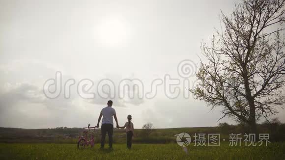 爸爸正在教女儿如何在日落时骑自行车在草地上。 幸福的父亲为女儿感到高兴