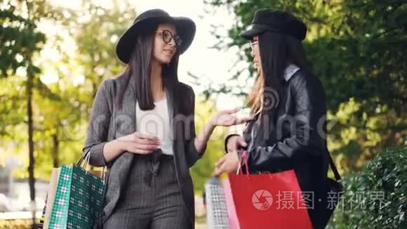 带着购物袋的快乐亚洲女孩正和她站在街上的朋友聊天，然后在街上展示她的购物品