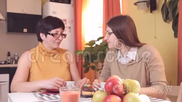 妈妈和带眼镜的小女儿在厨房喝茶或咖啡，吃蛋糕，说话，笑。 在前景中