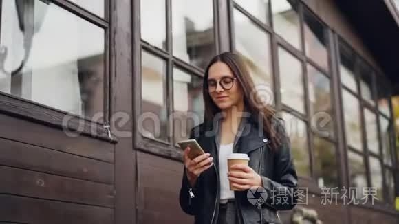 戴眼镜的可爱年轻女子用智能手机微笑着走在街上拿着外卖的慢动作