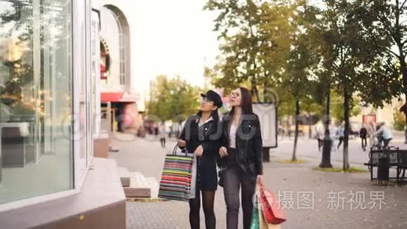 时尚的年轻女性朋友沿街走着，手里拿着纸袋购物，看着橱窗