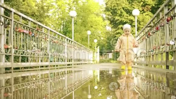 穿着橙色防水雨衣和橡胶雨靴的滑稽小女孩在雨后的水坑里慢动作