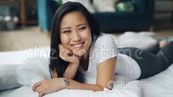 一幅美丽的亚洲女士躺在家里的床上，微笑着，然后笑着看着镜头。 放松，心情好