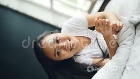 慢动作，美丽的亚洲黑发女人在床上翻滚，高兴地微笑着看着相机，享受着乐趣